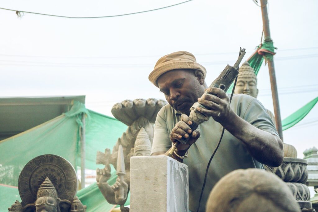 An artisan skillfully carves a stone statue in an outdoor workshop setting.